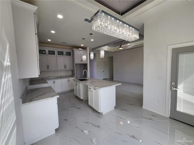 kitchen with a center island with sink, sink, hanging light fixtures, light stone countertops, and white cabinetry