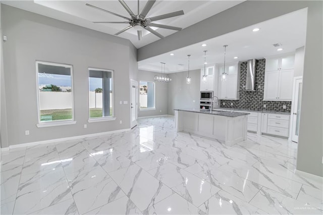 kitchen featuring built in microwave, wall chimney exhaust hood, pendant lighting, white cabinets, and ceiling fan with notable chandelier