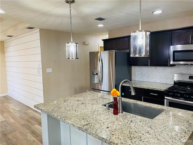 kitchen featuring sink, light stone counters, decorative light fixtures, stainless steel appliances, and a kitchen island with sink