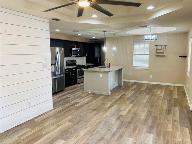 kitchen with appliances with stainless steel finishes, an island with sink, pendant lighting, a tray ceiling, and sink