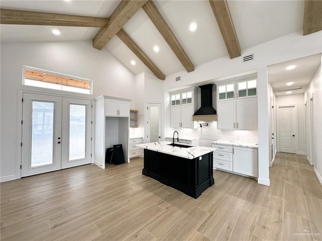 kitchen with french doors, high vaulted ceiling, white cabinets, a kitchen island with sink, and wall chimney range hood