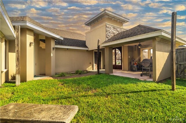 back of house at dusk featuring roof with shingles, stucco siding, a yard, and a patio area
