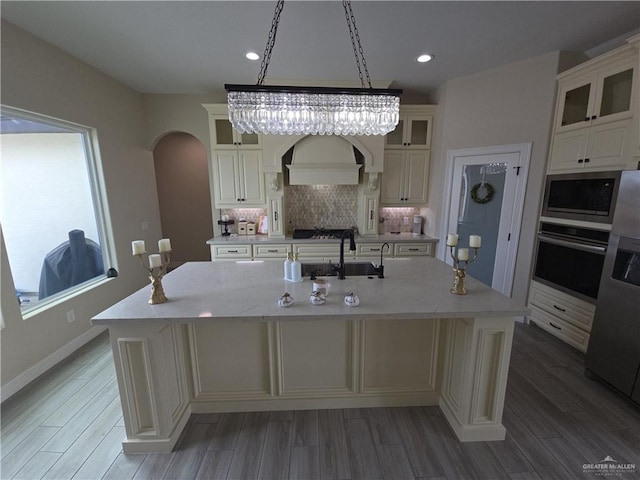 kitchen with premium range hood, stainless steel appliances, a kitchen island with sink, sink, and hanging light fixtures