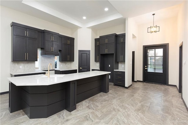 kitchen with sink, hanging light fixtures, tasteful backsplash, an island with sink, and a raised ceiling