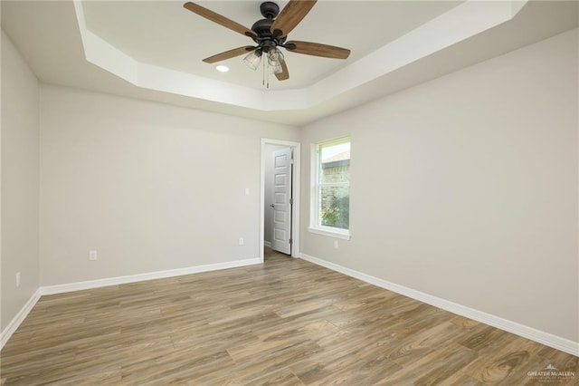 empty room with light wood-type flooring, a tray ceiling, baseboards, and ceiling fan