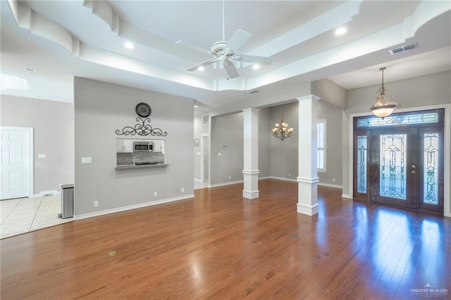 unfurnished living room featuring ornate columns, sink, a raised ceiling, ceiling fan with notable chandelier, and light wood-type flooring