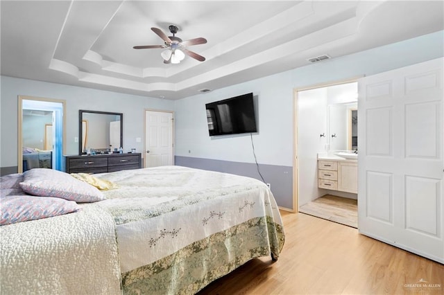 bedroom with light wood-type flooring, ceiling fan, a raised ceiling, and ensuite bath