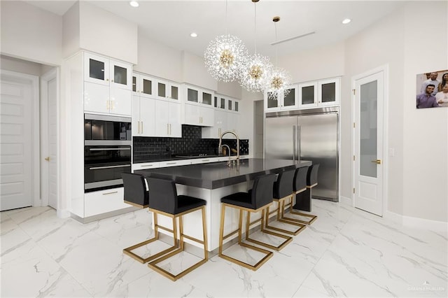 kitchen with built in appliances, dark countertops, an island with sink, and glass insert cabinets