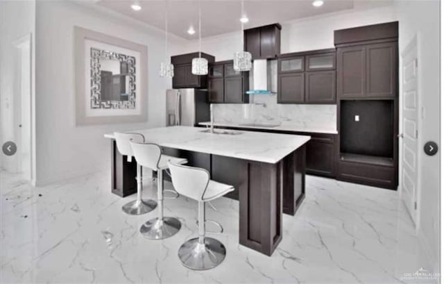 kitchen with a center island with sink, stainless steel fridge, decorative backsplash, hanging light fixtures, and wall chimney range hood