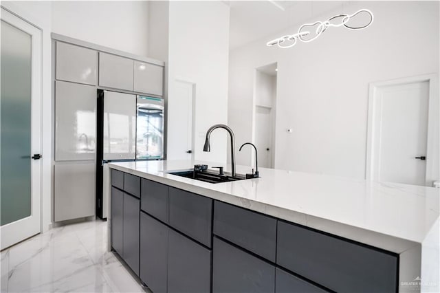 kitchen with light stone countertops, stainless steel fridge, gray cabinetry, a kitchen island with sink, and sink