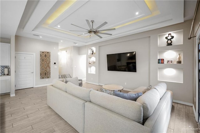 living room featuring built in shelves, a tray ceiling, light wood finished floors, and a ceiling fan