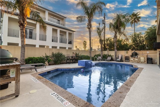 pool at dusk featuring pool water feature, an outdoor kitchen, a patio, and grilling area