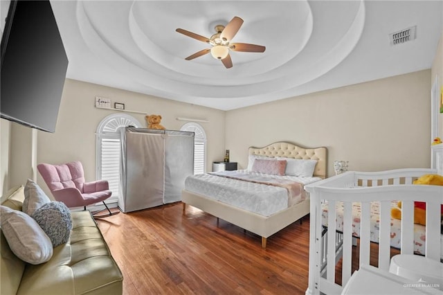 bedroom featuring a raised ceiling, wood finished floors, visible vents, and a ceiling fan