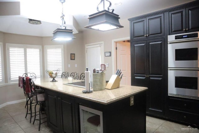 kitchen with light tile patterned flooring, double oven, decorative light fixtures, light stone counters, and a center island with sink