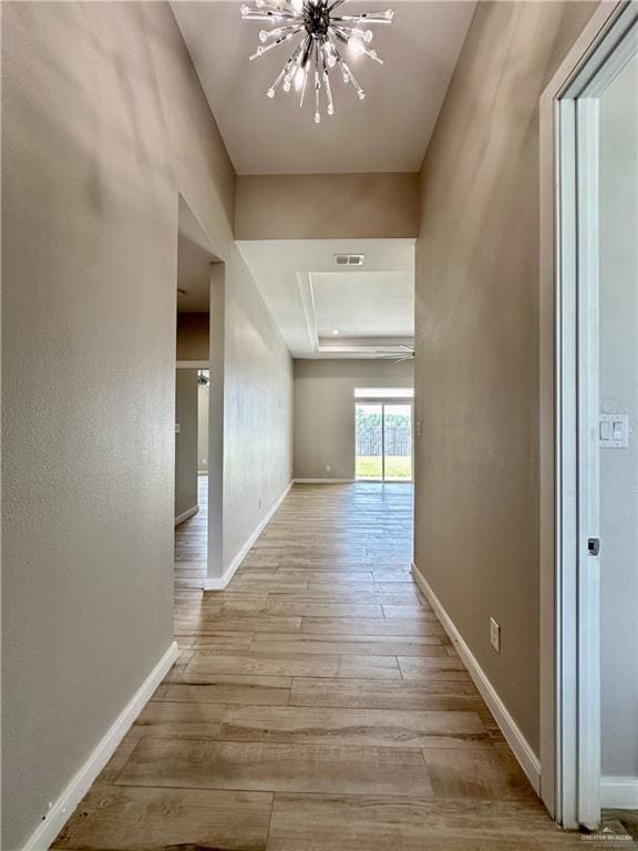 hallway with light wood-type flooring, a tray ceiling, and an inviting chandelier