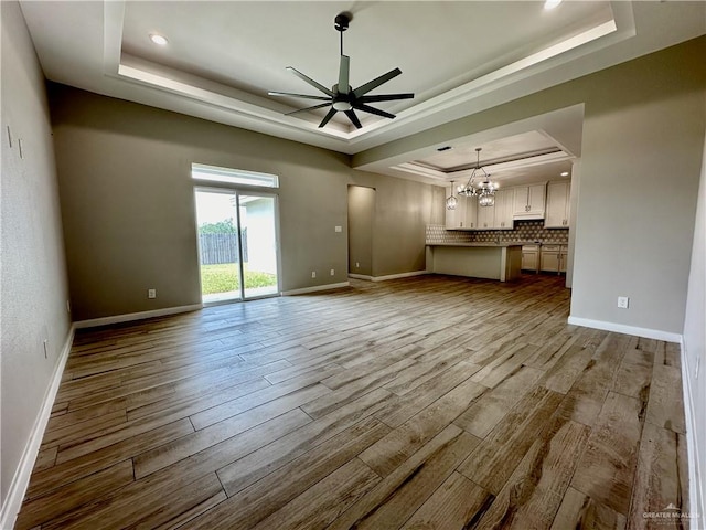 unfurnished living room with ceiling fan with notable chandelier, light wood-type flooring, and a tray ceiling