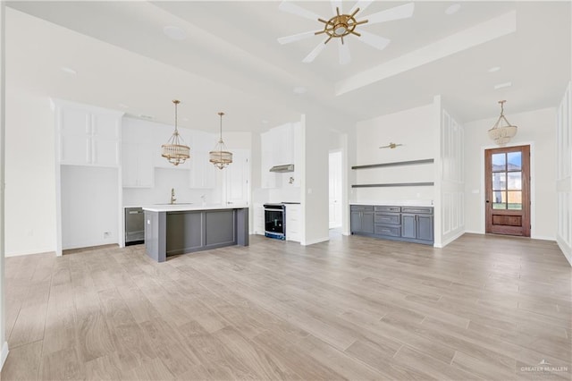 unfurnished living room with a sink, a ceiling fan, baseboards, light wood-style floors, and a tray ceiling