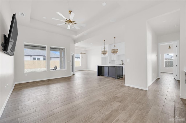 unfurnished living room featuring visible vents, a tray ceiling, a healthy amount of sunlight, light wood-style floors, and ceiling fan with notable chandelier