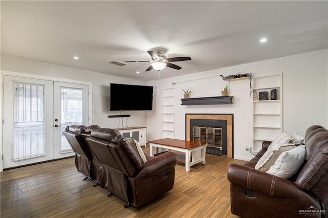 living room featuring hardwood / wood-style flooring, built in features, ceiling fan, a tiled fireplace, and french doors