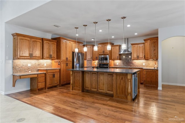 kitchen with appliances with stainless steel finishes, light wood-type flooring, wall chimney exhaust hood, a kitchen island with sink, and decorative light fixtures