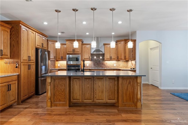 kitchen featuring hardwood / wood-style floors, a kitchen island with sink, hanging light fixtures, wall chimney exhaust hood, and appliances with stainless steel finishes