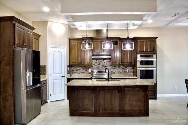 kitchen with light stone counters, stainless steel appliances, sink, decorative light fixtures, and a center island with sink