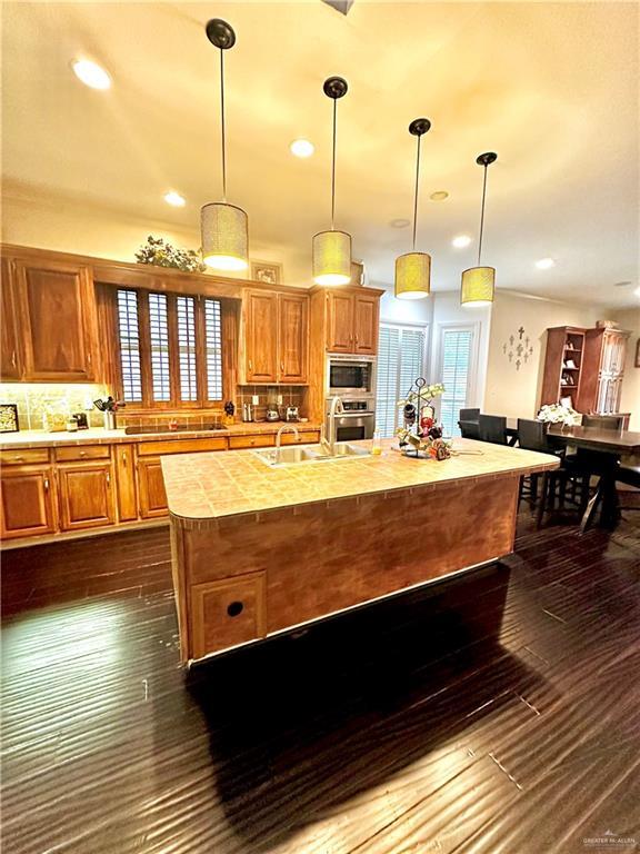 kitchen featuring pendant lighting, dark wood-type flooring, backsplash, stainless steel appliances, and an island with sink