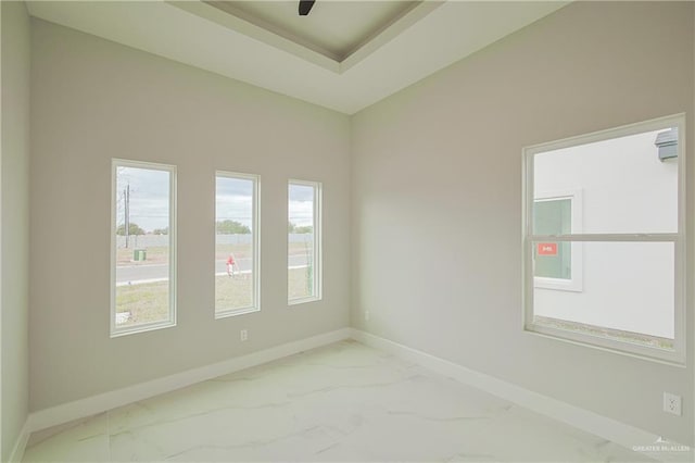 empty room with ceiling fan, marble finish floor, a tray ceiling, and baseboards