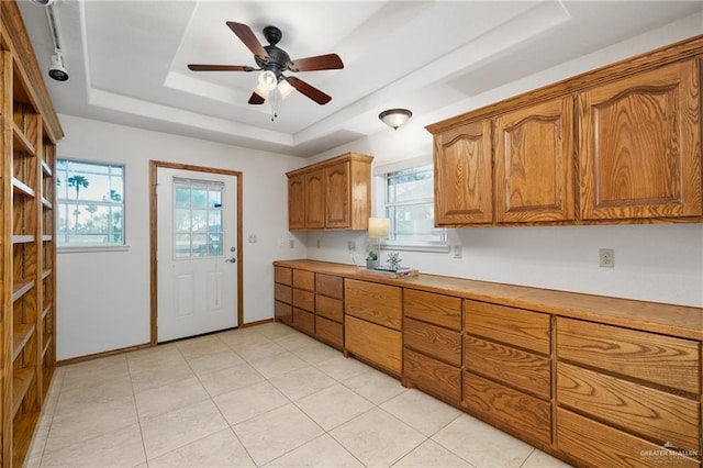 kitchen with baseboards, brown cabinetry, ceiling fan, a tray ceiling, and light countertops