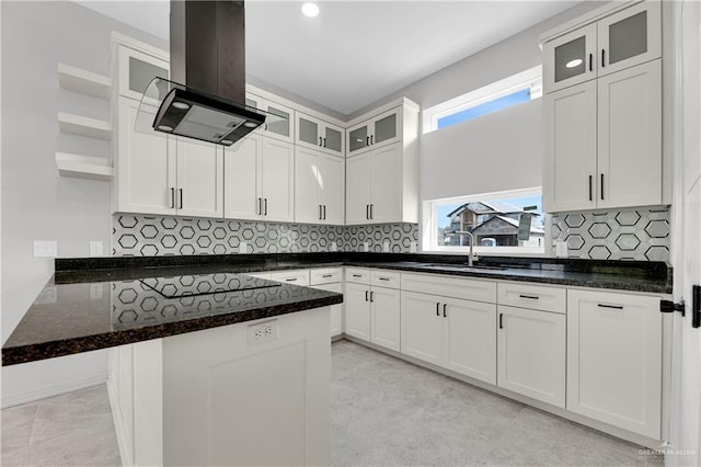 kitchen with a sink, island exhaust hood, dark stone counters, backsplash, and white cabinets
