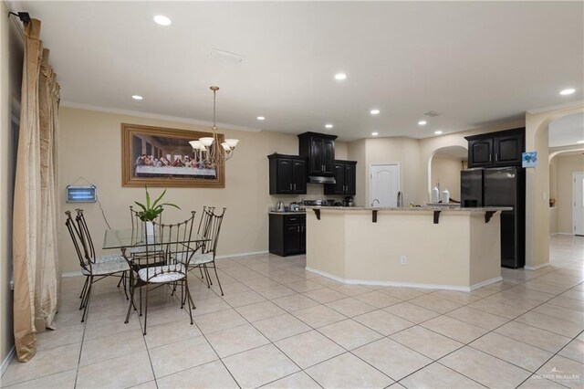 kitchen with black refrigerator, crown molding, light tile patterned floors, an island with sink, and a notable chandelier