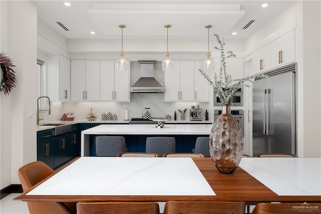 kitchen featuring wall chimney range hood, pendant lighting, a center island with sink, white cabinets, and appliances with stainless steel finishes