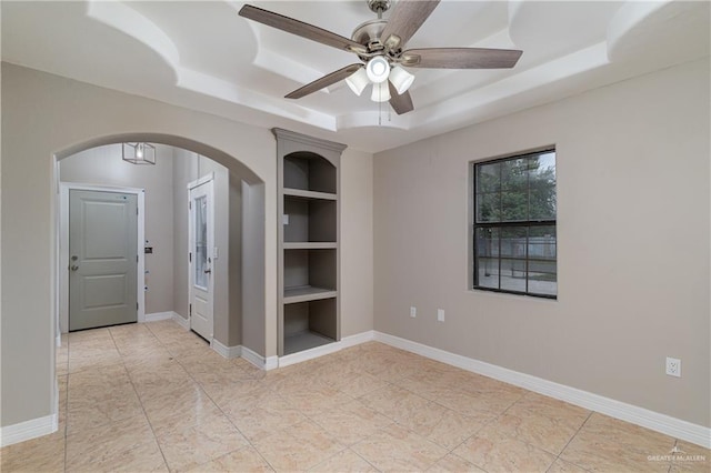 unfurnished room with a raised ceiling, built in shelves, and ceiling fan