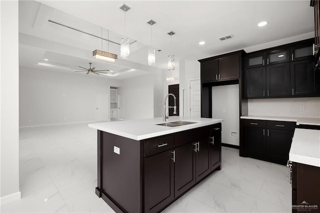 kitchen with ceiling fan, sink, pendant lighting, a tray ceiling, and a kitchen island with sink