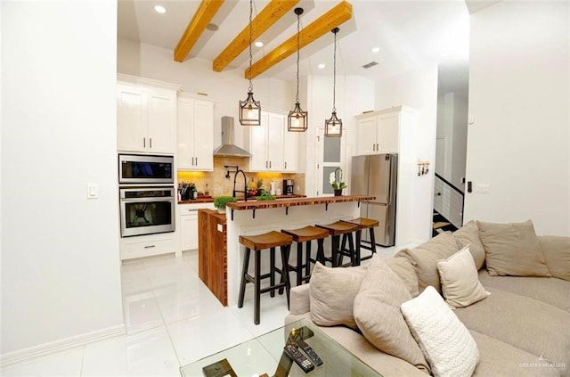 kitchen featuring pendant lighting, wall chimney exhaust hood, appliances with stainless steel finishes, beamed ceiling, and white cabinetry