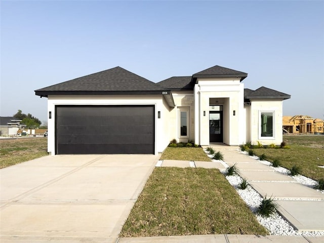 prairie-style home with a front yard, driveway, an attached garage, a shingled roof, and stucco siding