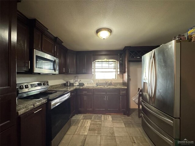 kitchen featuring dark brown cabinetry, a sink, a textured ceiling, dark stone countertops, and stainless steel appliances