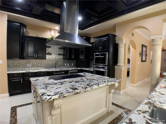 kitchen with island exhaust hood, black electric cooktop, decorative columns, dark cabinetry, and arched walkways
