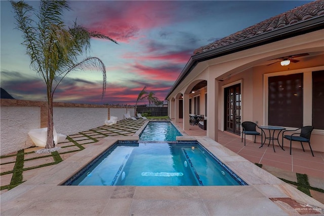 pool at dusk featuring a water view, ceiling fan, and a patio area