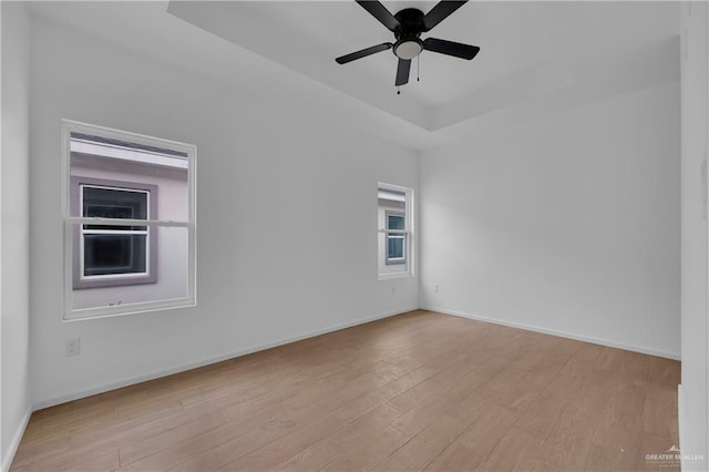 empty room with a ceiling fan, a tray ceiling, baseboards, and light wood finished floors