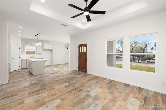 foyer with a raised ceiling, ceiling fan with notable chandelier, a wealth of natural light, and light wood-type flooring