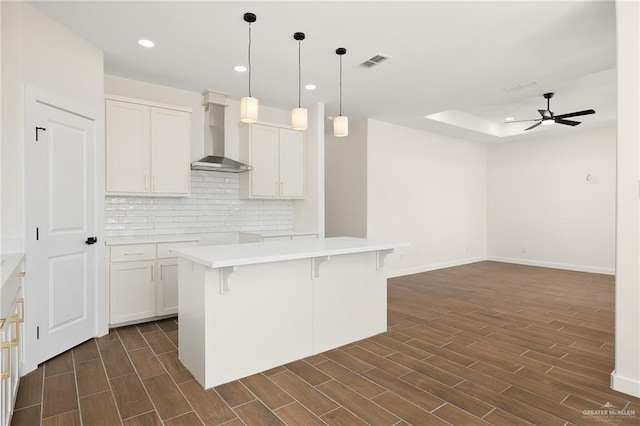 kitchen featuring a kitchen island with sink, wall chimney range hood, white cabinets, and decorative light fixtures