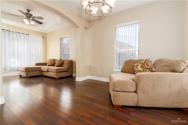 living room with arched walkways, dark wood-style flooring, a tray ceiling, baseboards, and ceiling fan with notable chandelier