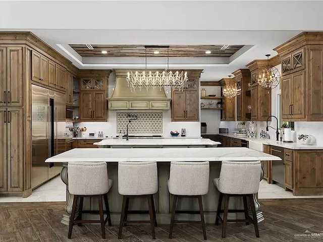 kitchen with an island with sink, a tray ceiling, stainless steel built in refrigerator, premium range hood, and open shelves