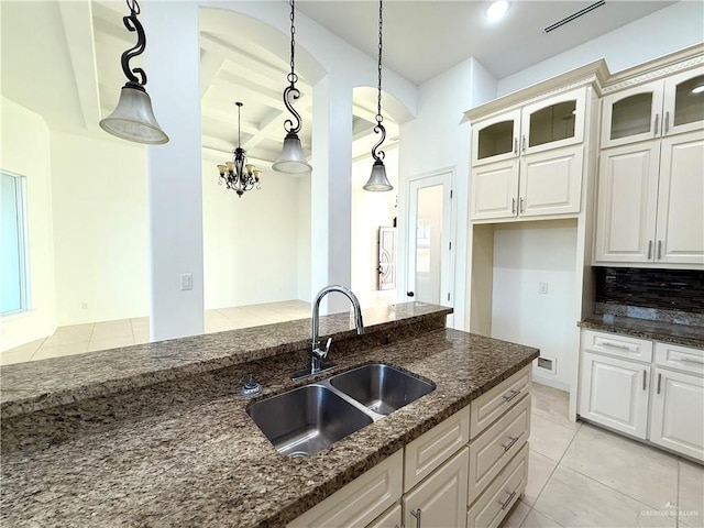 kitchen with light tile patterned flooring, coffered ceiling, a sink, dark stone countertops, and glass insert cabinets