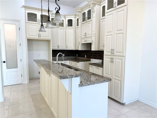 kitchen featuring decorative light fixtures, a center island with sink, light tile patterned floors, glass insert cabinets, and dark stone counters