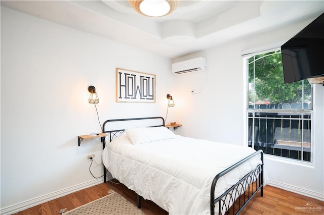 bedroom featuring a wall mounted AC, wood finished floors, a raised ceiling, and baseboards