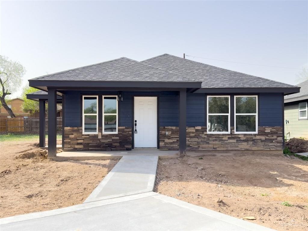 view of front of house featuring stone siding, a shingled roof, fence, and a porch