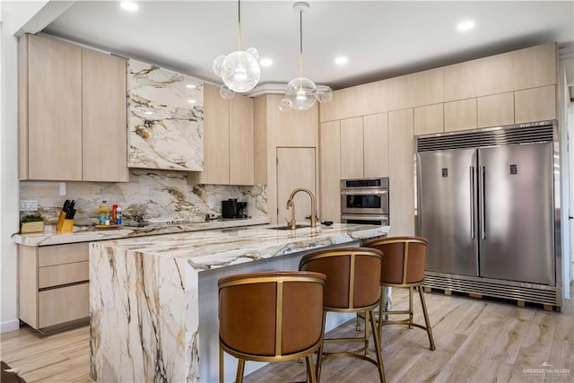 kitchen with light brown cabinetry, a center island with sink, stainless steel appliances, and light hardwood / wood-style flooring