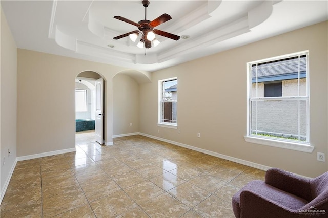 empty room featuring arched walkways, a raised ceiling, a ceiling fan, and baseboards